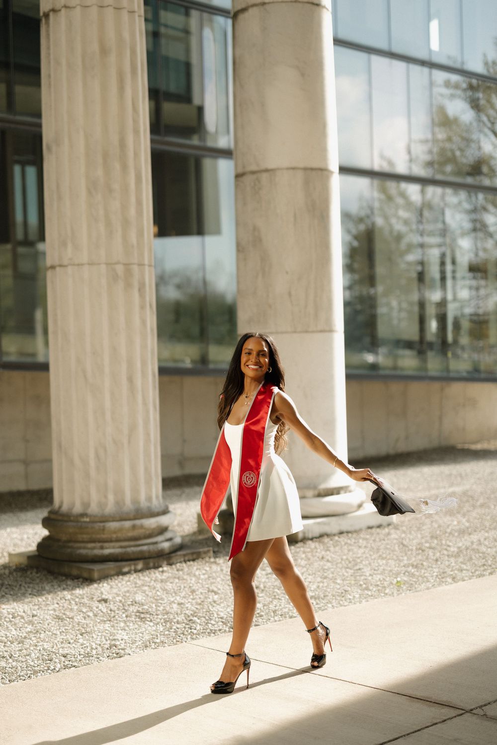 Confident female graduate in white dress and red stole walks past marble columns on campus.