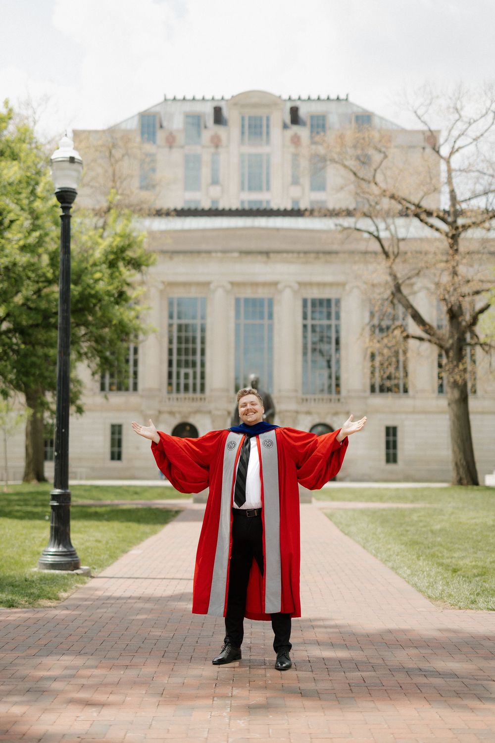 Graduate in red doctoral regalia stands with arms outstretched in front of a classic university building.