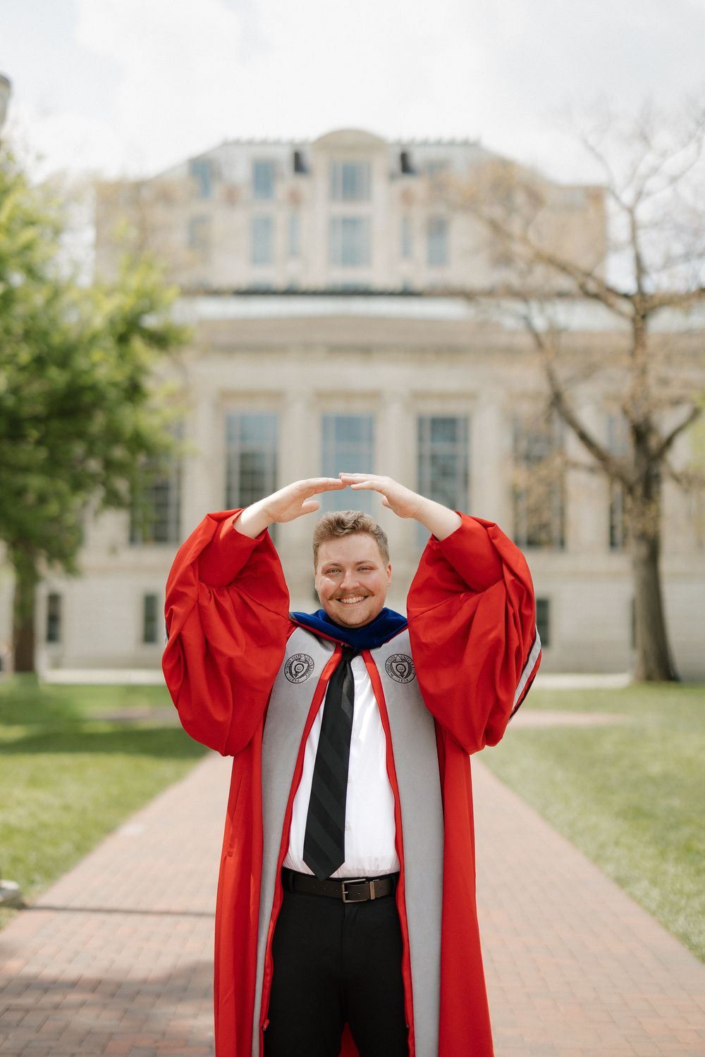 Cheerful doctoral graduate in red regalia forms letter O with arms in front of a university building.