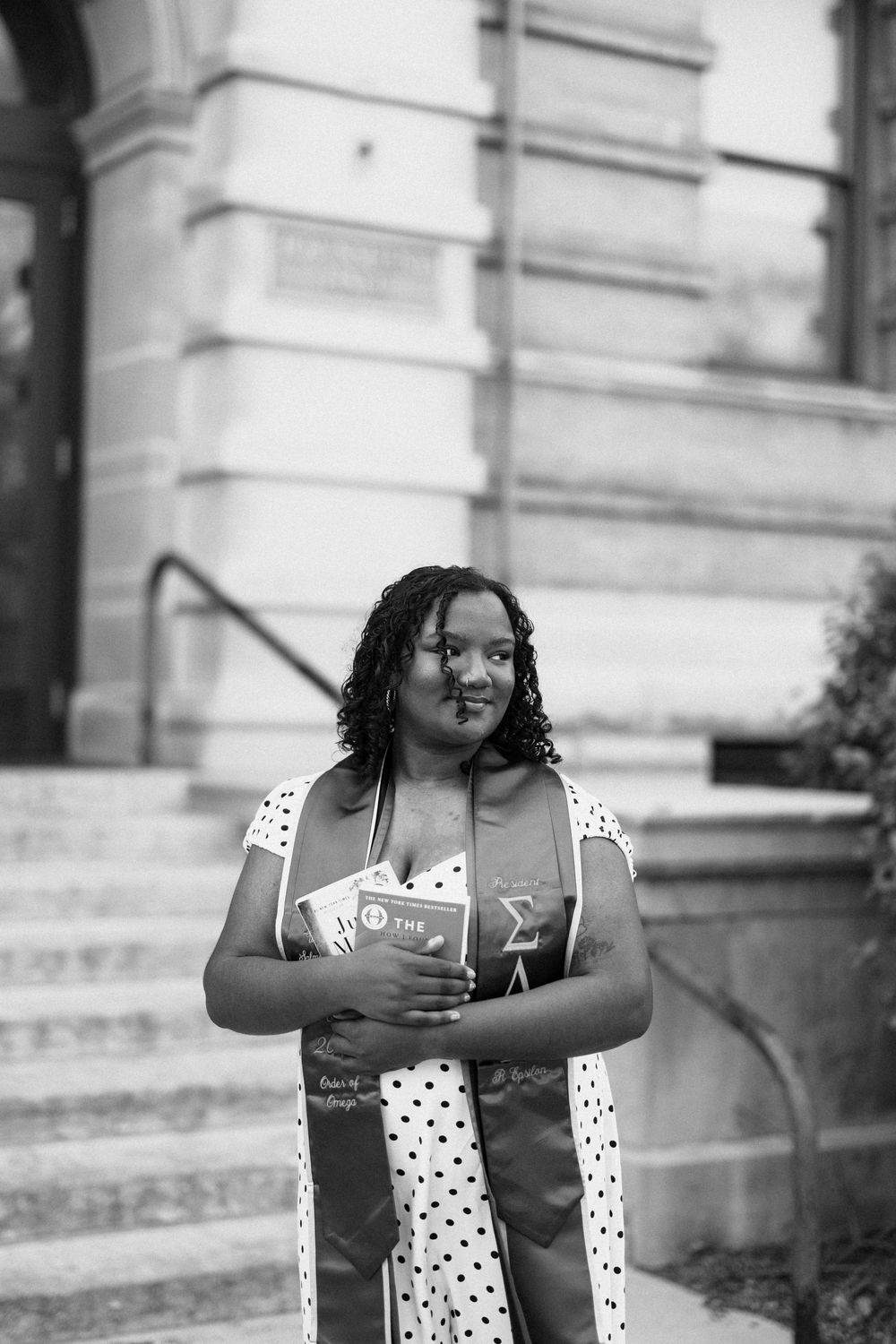Black and white portrait of a graduate in polka dot dress holding books outside a historic university building.
