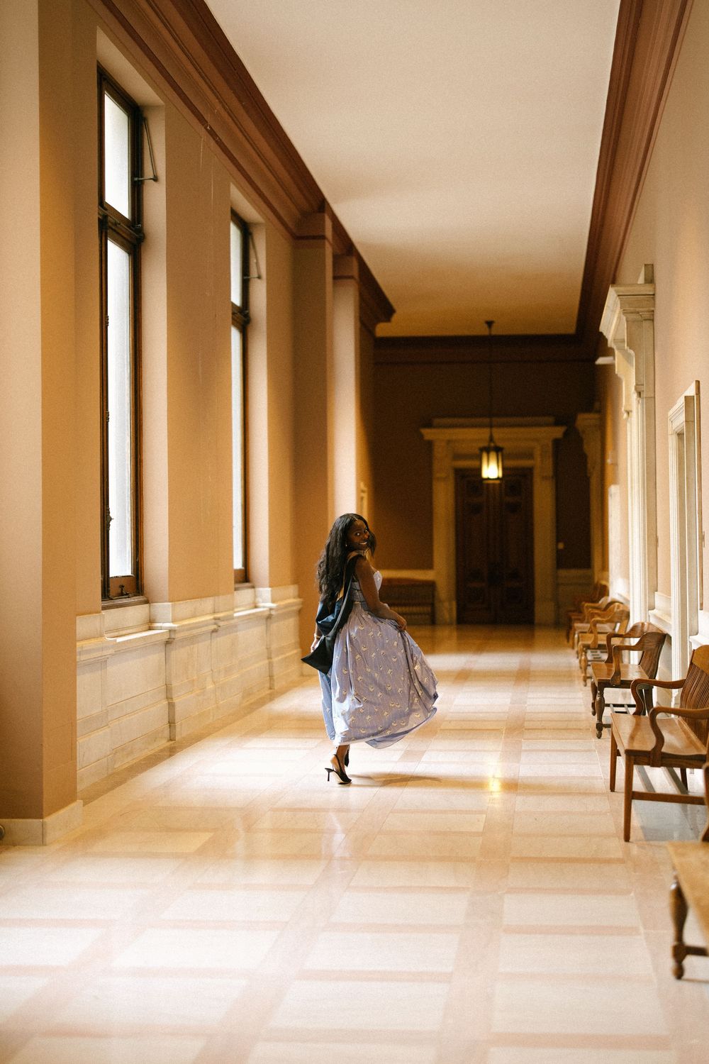 Woman in silver dress and leather jacket walks down sunlit marble corridor of grand building.