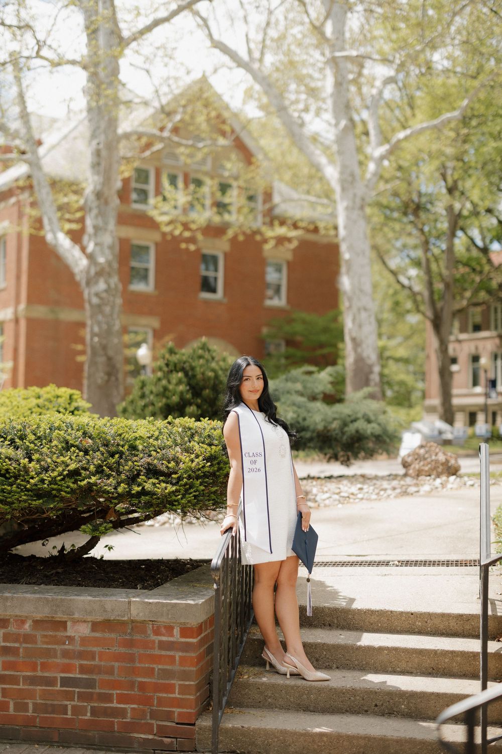 Elegant graduate in white dress holds diploma cover while posing on steps outside a brick university building.