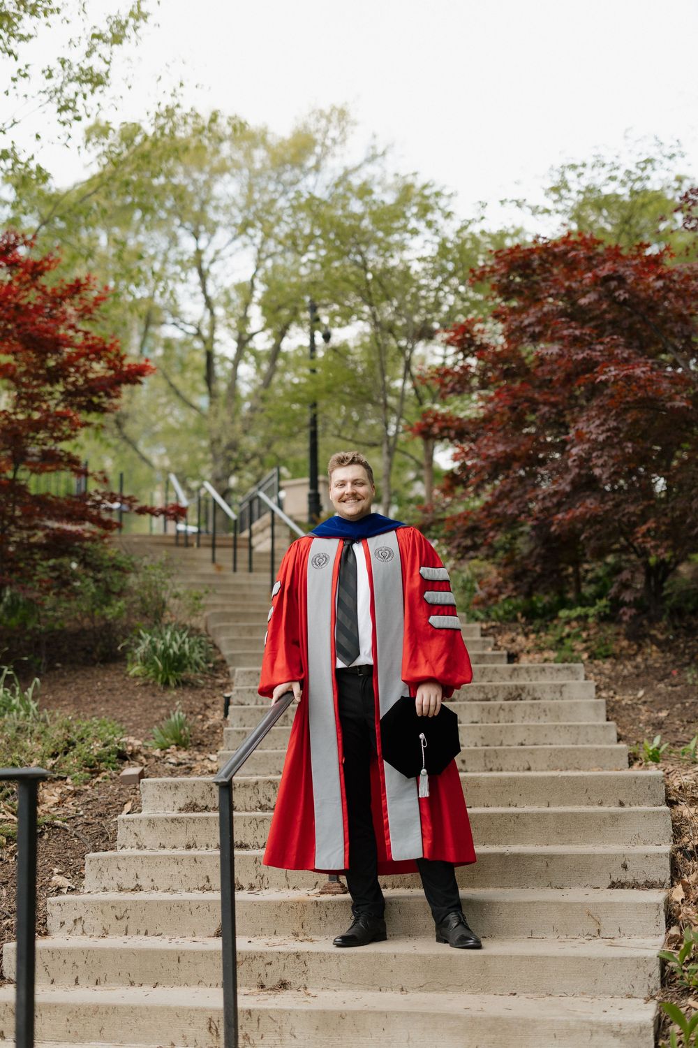 Smiling PhD graduate in red doctoral gown stands on stone steps surrounded by colorful autumn trees.