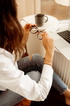 Person wearing white shirt and jeans working by laptop with coffee mug and glasses.