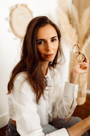 Contemporary portrait of someone in a white button-down shirt holding eyeglasses in a warm-toned indoor setting.