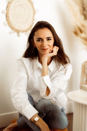 Lifestyle portrait of a woman in a white button down shirt and jeans sitting casually in a bright interior setting.