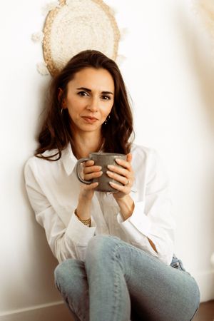 Portrait style photo of someone sitting with mug wearing white shirt and blue jeans.