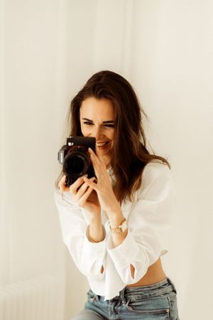 Photographer in white shirt and jeans laughing while holding camera against light background.