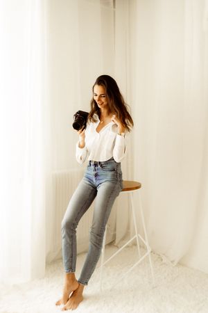 A photographer in a white shirt and light jeans works barefoot while holding a professional camera in a bright studio setting.