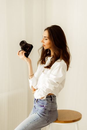 Person in white button down shirt and jeans examining camera near window.