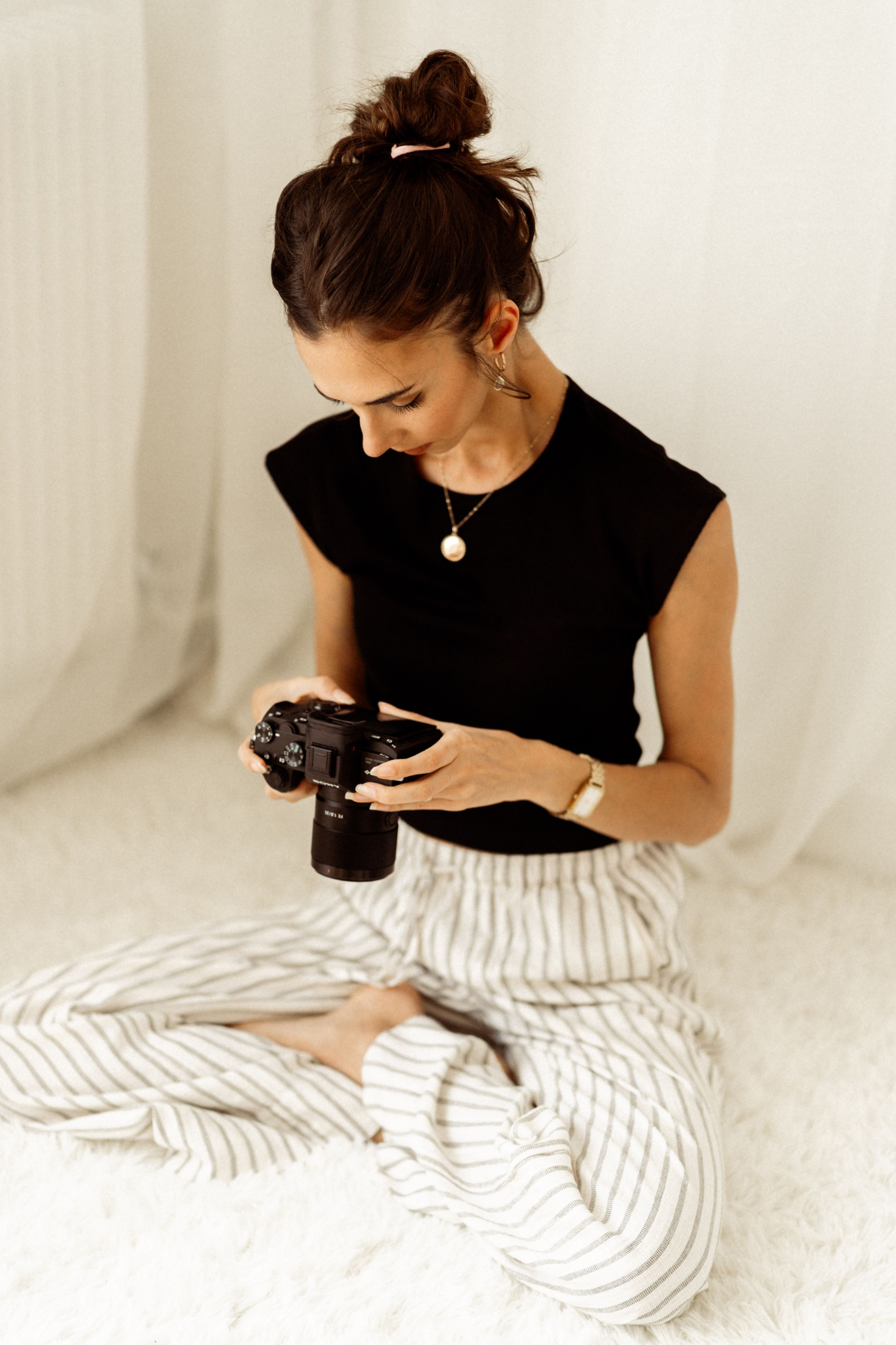 Person in black top and striped pants sits cross-legged while looking down at a camera.