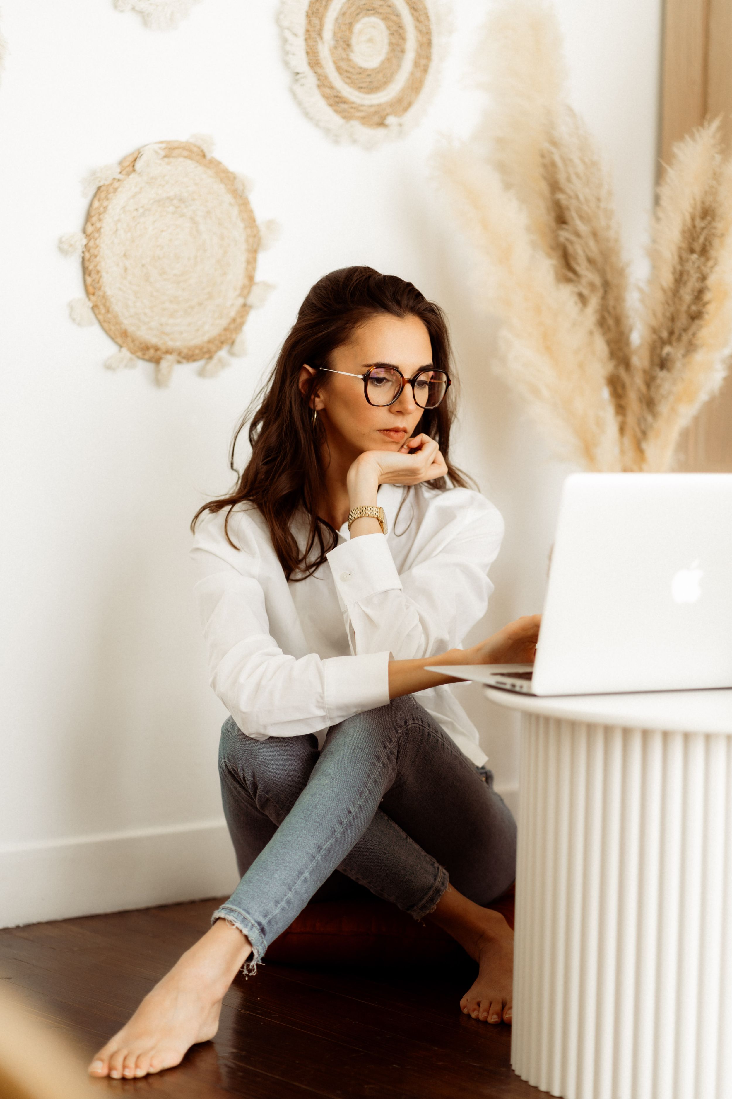 Person in glasses working on laptop while sitting on floor with pampas grass decor and circular wall art in background.