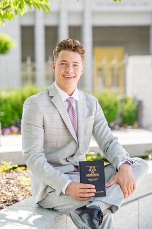 A person in a light gray suit with a pink tie holding The Book of Mormon while sitting outside a temple.