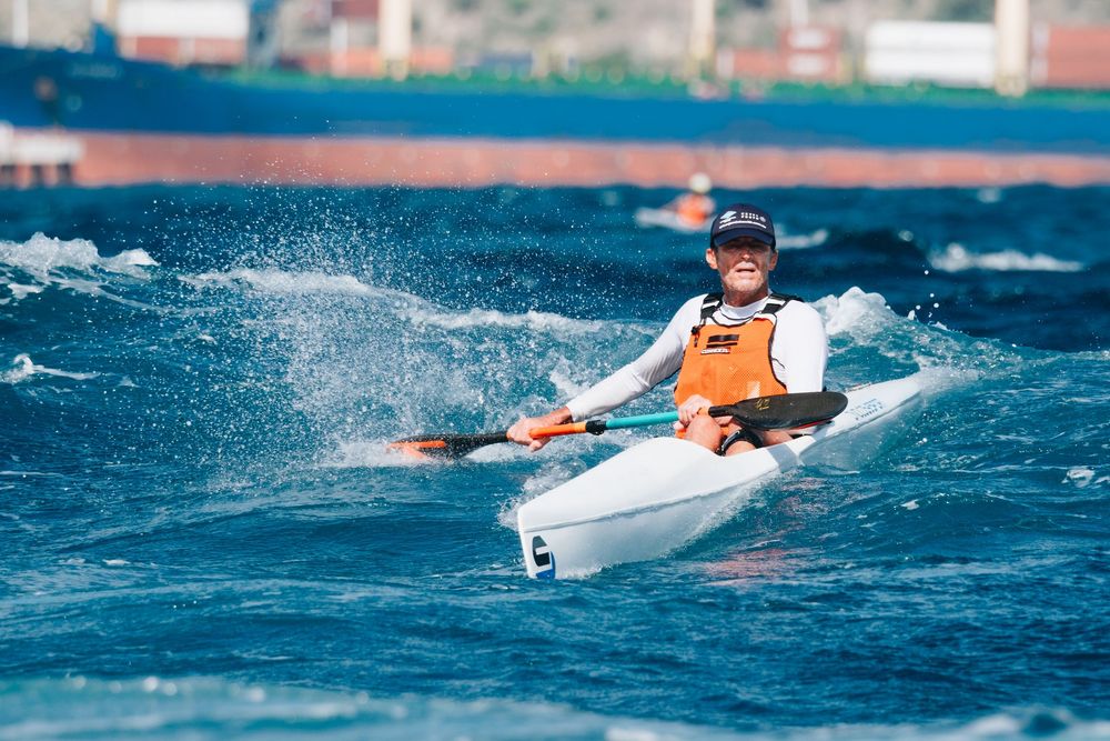 A kayaker in an orange life vest paddles through blue ocean waves on a sunny day.