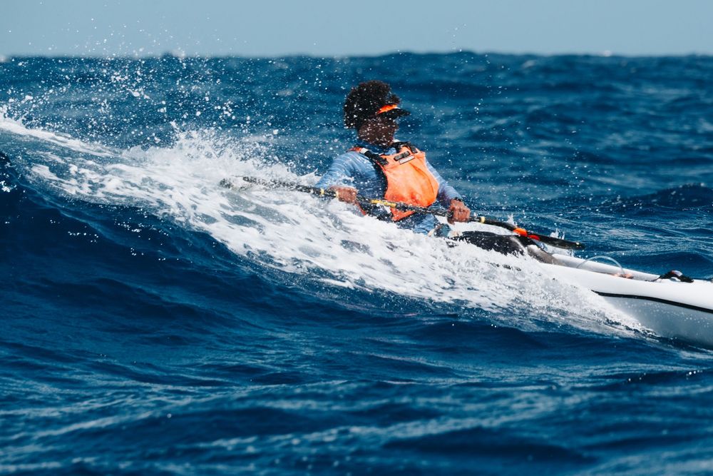 Sea kayaker paddling through blue ocean waves with white spray trailing behind the vessel.