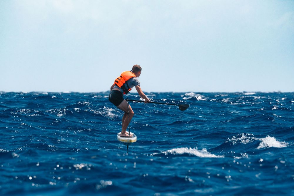 A surfer in an orange life vest rides a hydrofoil board across deep blue ocean waves.