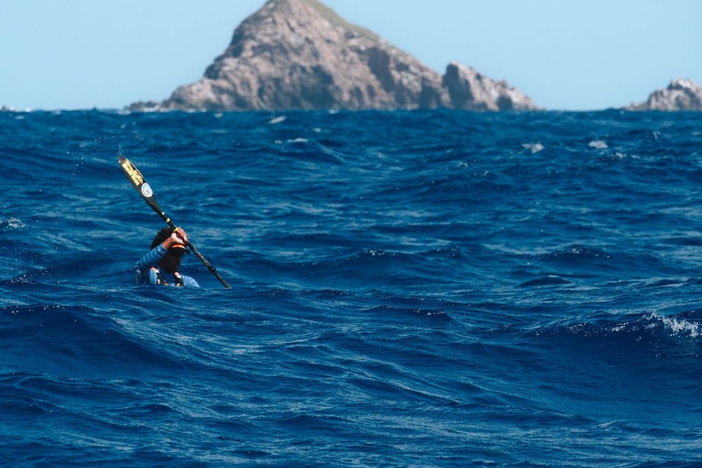A seabird spreads its wings while flying over deep blue ocean waves with a rocky island mountain in the background.