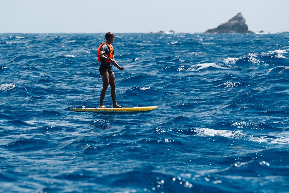 Paddle boarder in orange lifejacket rides across choppy blue ocean waves with distant island visible on horizon.