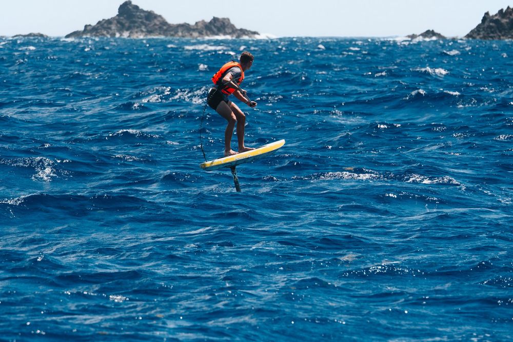 Surfer rides a hydrofoil board across deep blue choppy ocean waters with rocky islands in the background.
