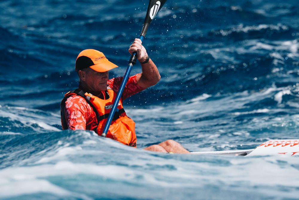 Kayaker in orange life vest and cap paddles through blue ocean waves during water sports adventure.