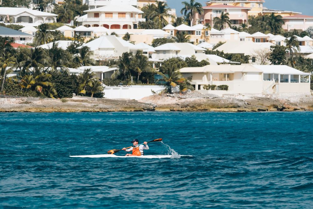 Two kayakers paddle in turquoise waters near a coastal residential area with white buildings and palm trees.