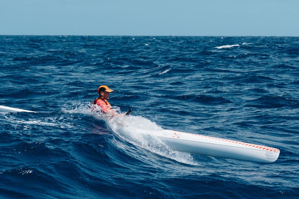 Person riding a white surfboard or paddleboard on choppy blue ocean waves on a bright sunny day.