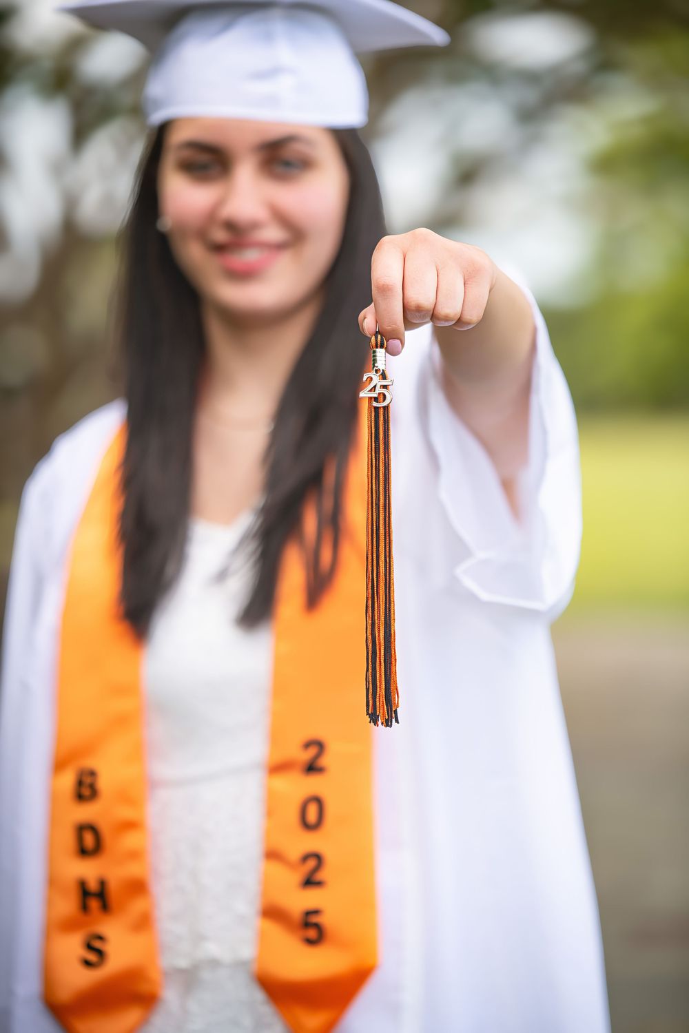 A graduate holds up their graduation tassel while wearing orange stole and white cap and gown.