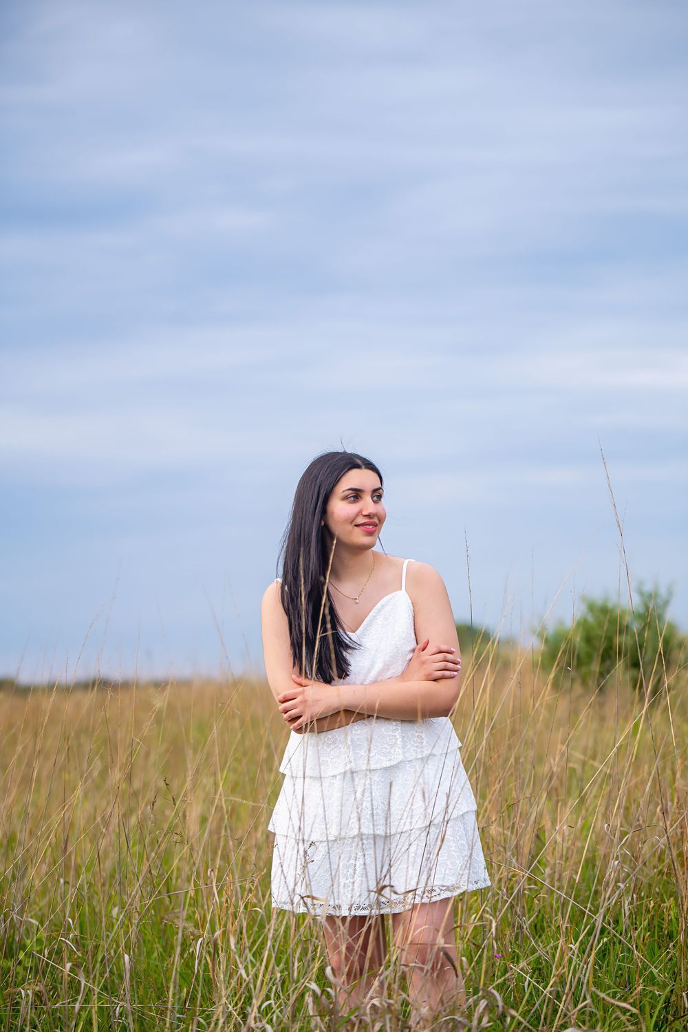 A summer portrait of a young woman in a white sundress standing in a tall grass field against a blue sky.