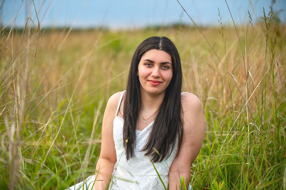 A woman in a white dress poses in a grassy meadow on a sunny day.
