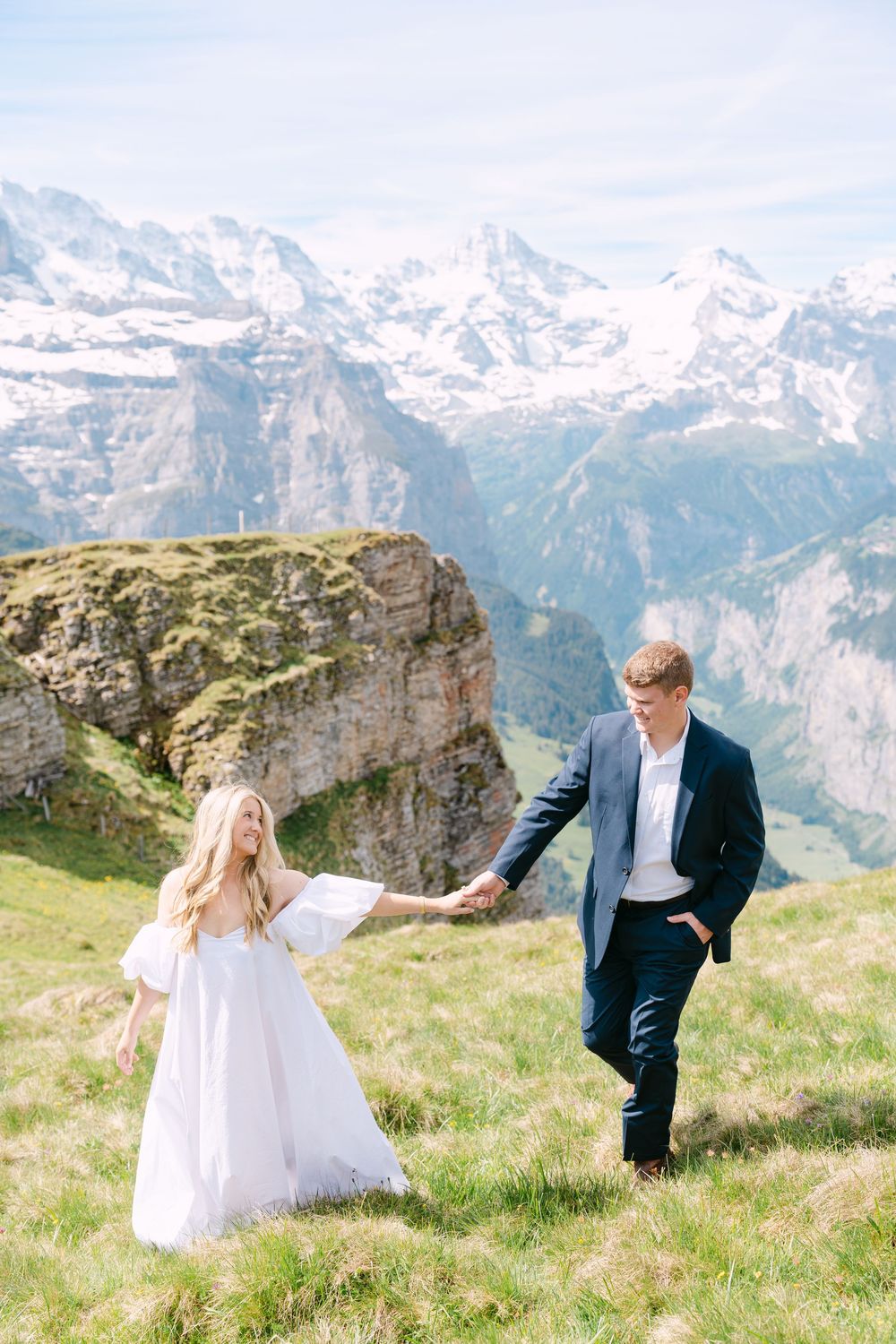 Couple holding hands on alpine meadow with snow-capped Swiss mountains backdrop, bride in white dress, groom in navy suit.