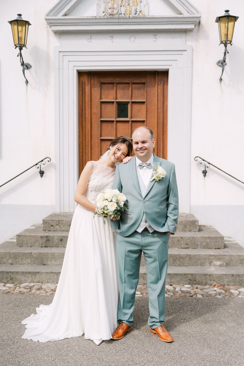 A wedding couple poses outside a white church building with ornate lanterns and wooden doors showing joy and celebration.