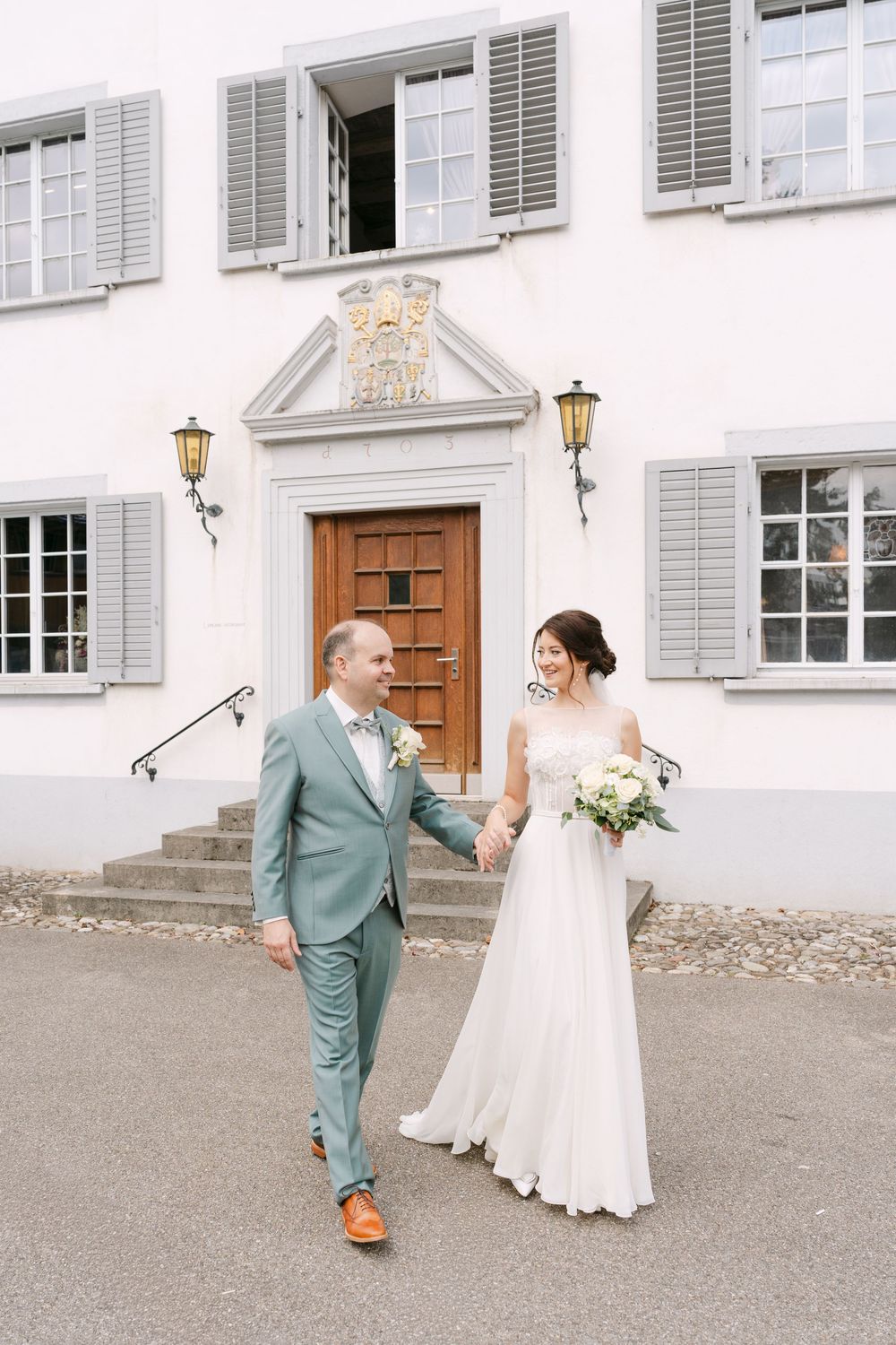 A romantic wedding couple shares a tender moment in front of a classic white building with gray shutters.
