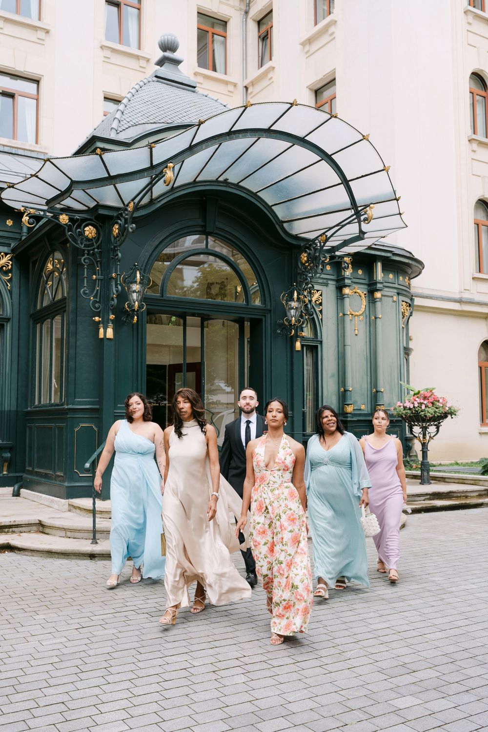 A group in pastel and floral dresses walking together under an elegant glass and metal archway entrance in Lausanne.