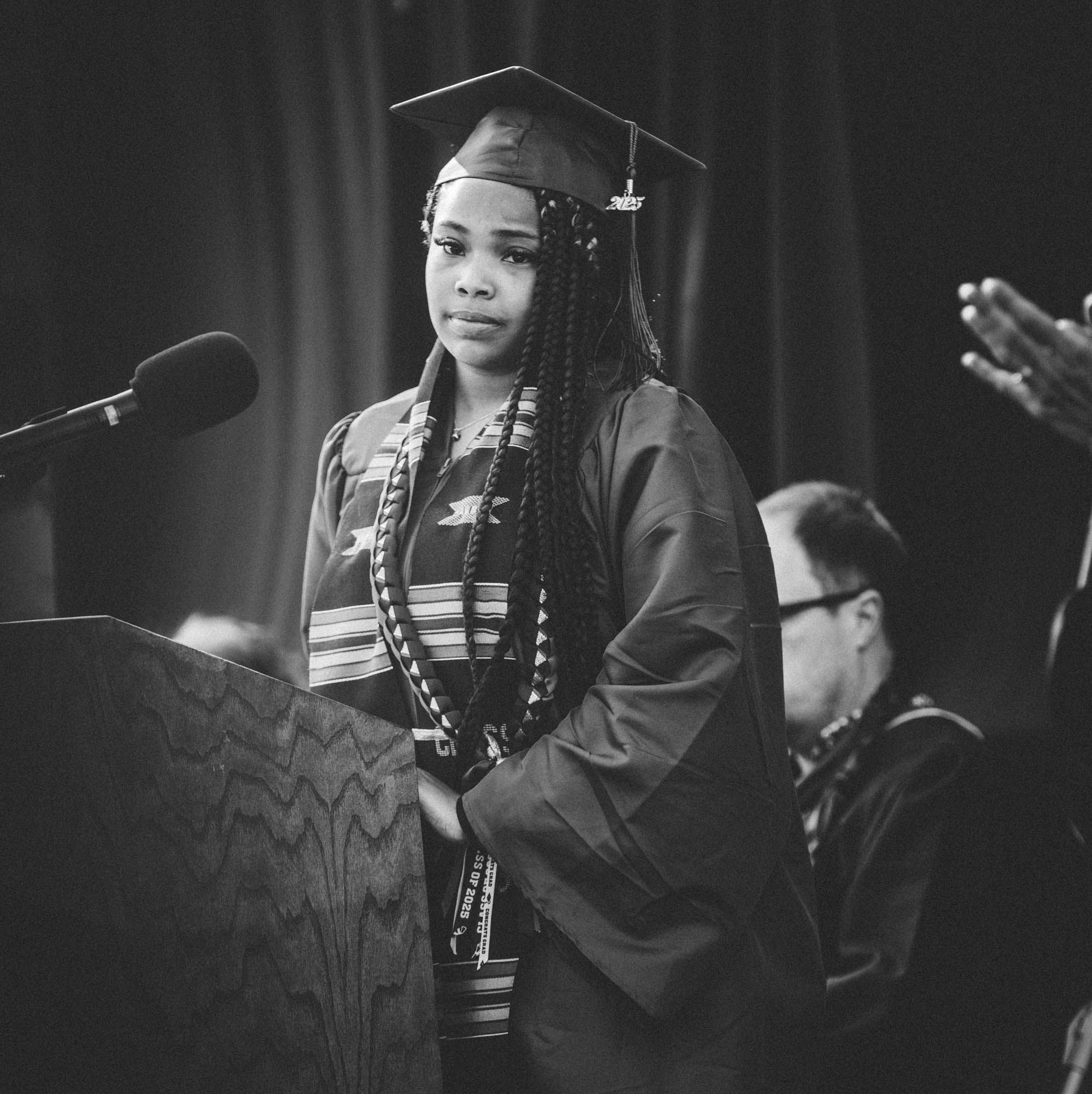 A graduate in academic regalia addresses an audience from a podium during a commencement ceremony in black and white.