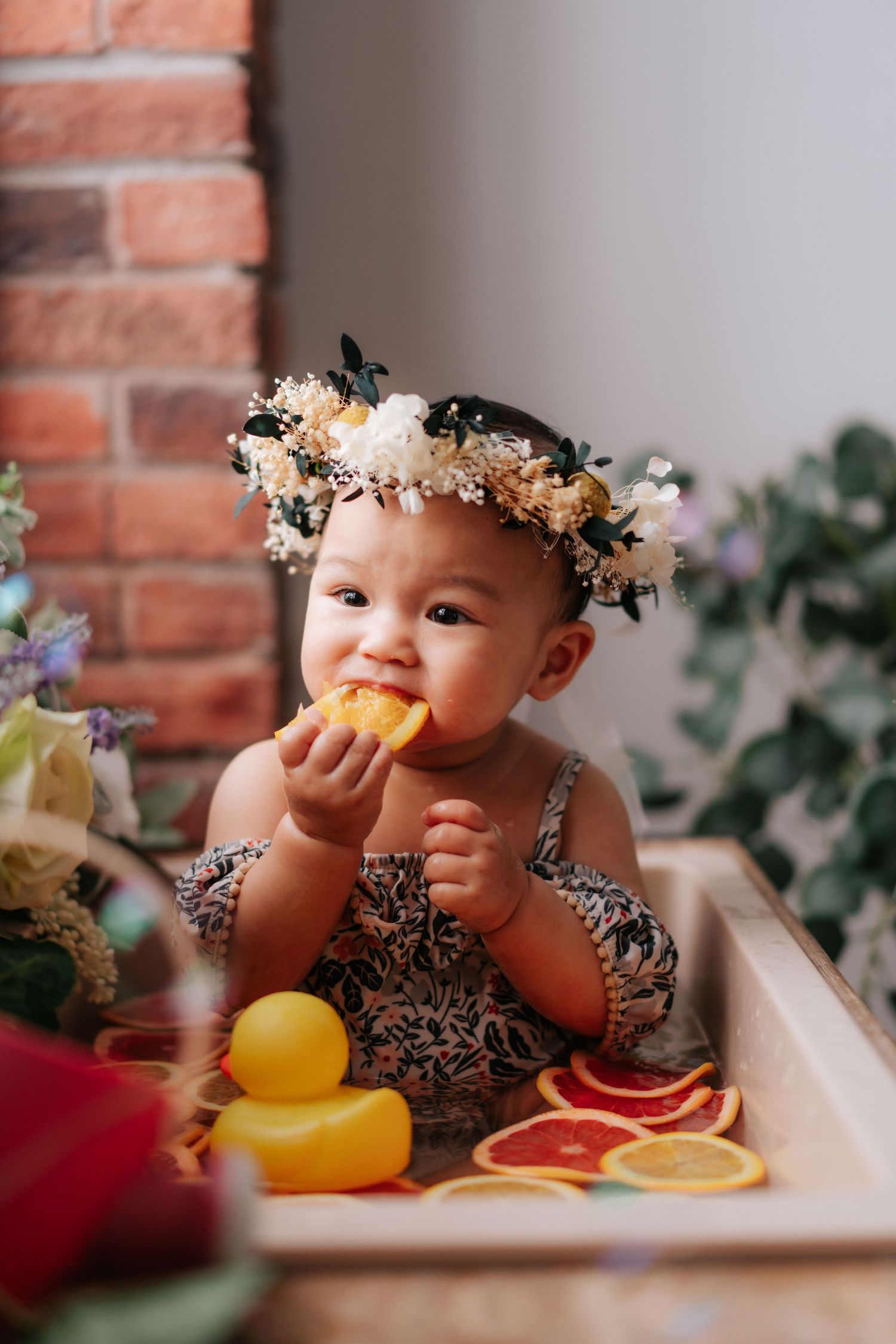 Baby wearing floral crown eating citrus fruit with decorative duck.