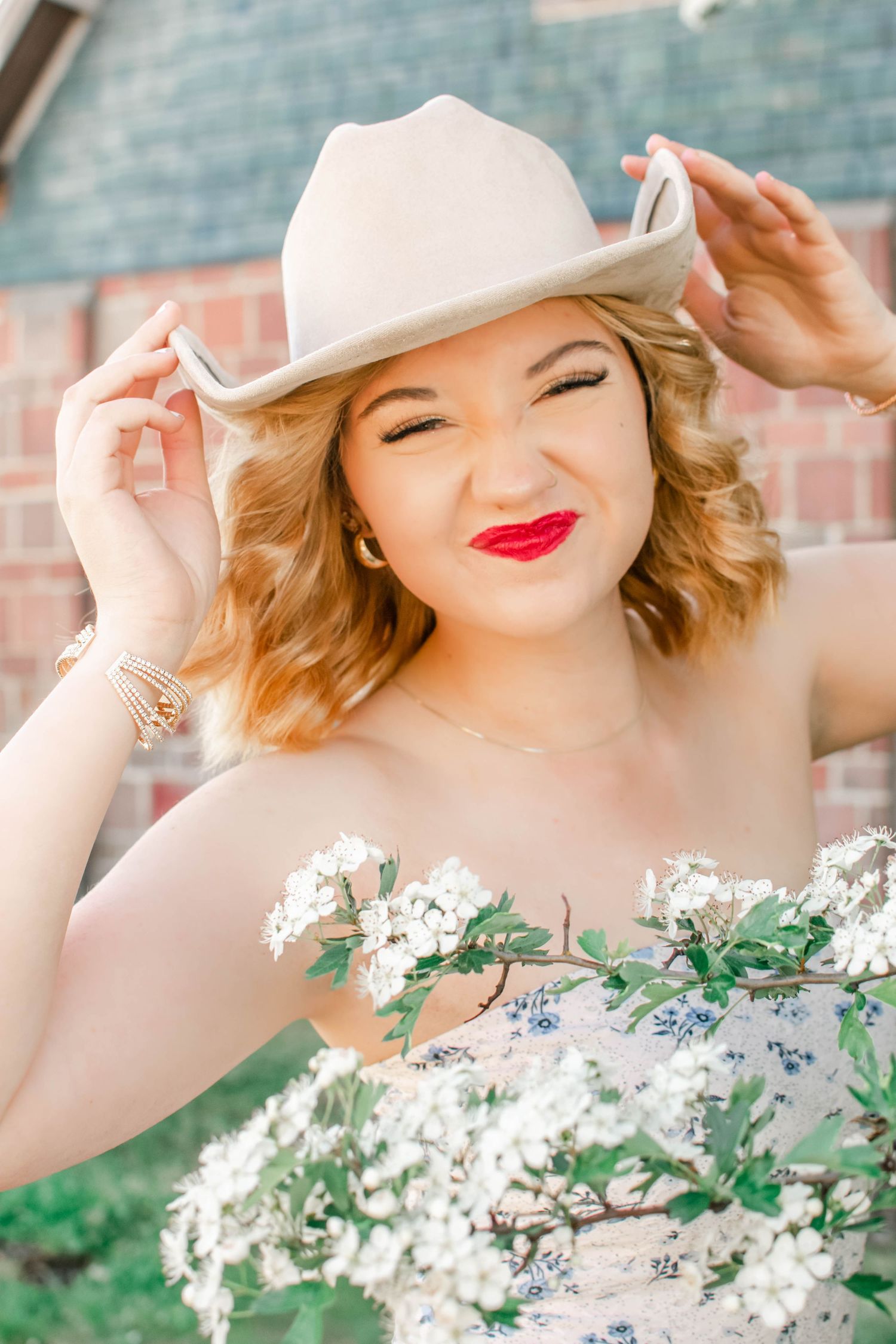 Elegant outdoor portrait with white hat and floral dress against rustic brick setting.