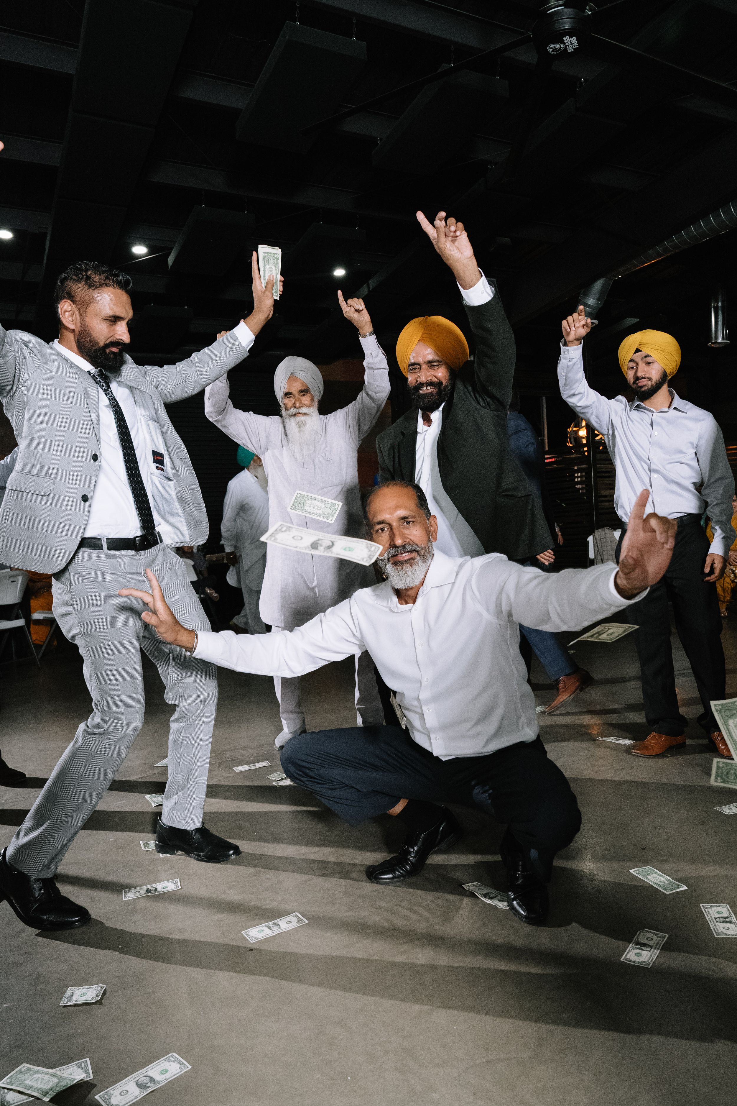 Group of Sikh businessmen celebrating with money raining down in a dark room, wearing suits and colorful turbans.