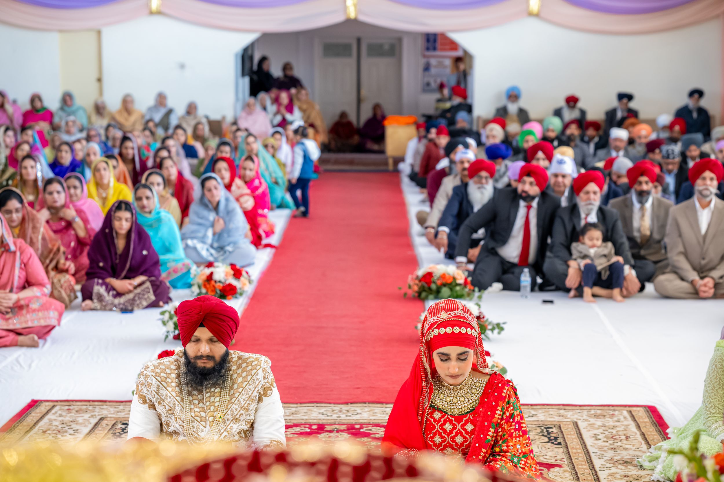 Traditional Sikh wedding ceremony with couples seated on floor with red carpet and guests in colorful attire in background.