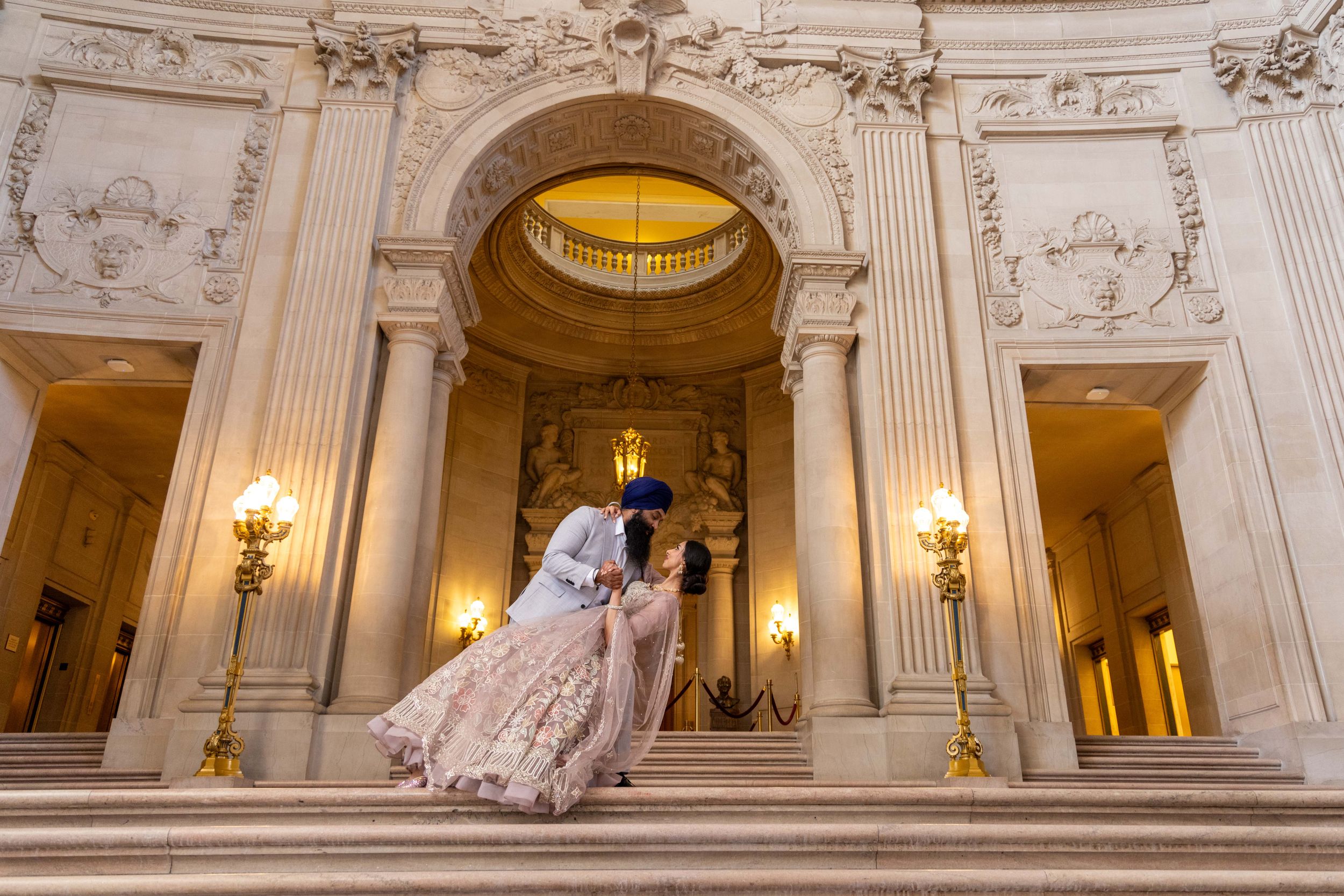 Two figures in elegant wedding attire embrace on grand marble steps inside an ornate building with glowing wall sconces.