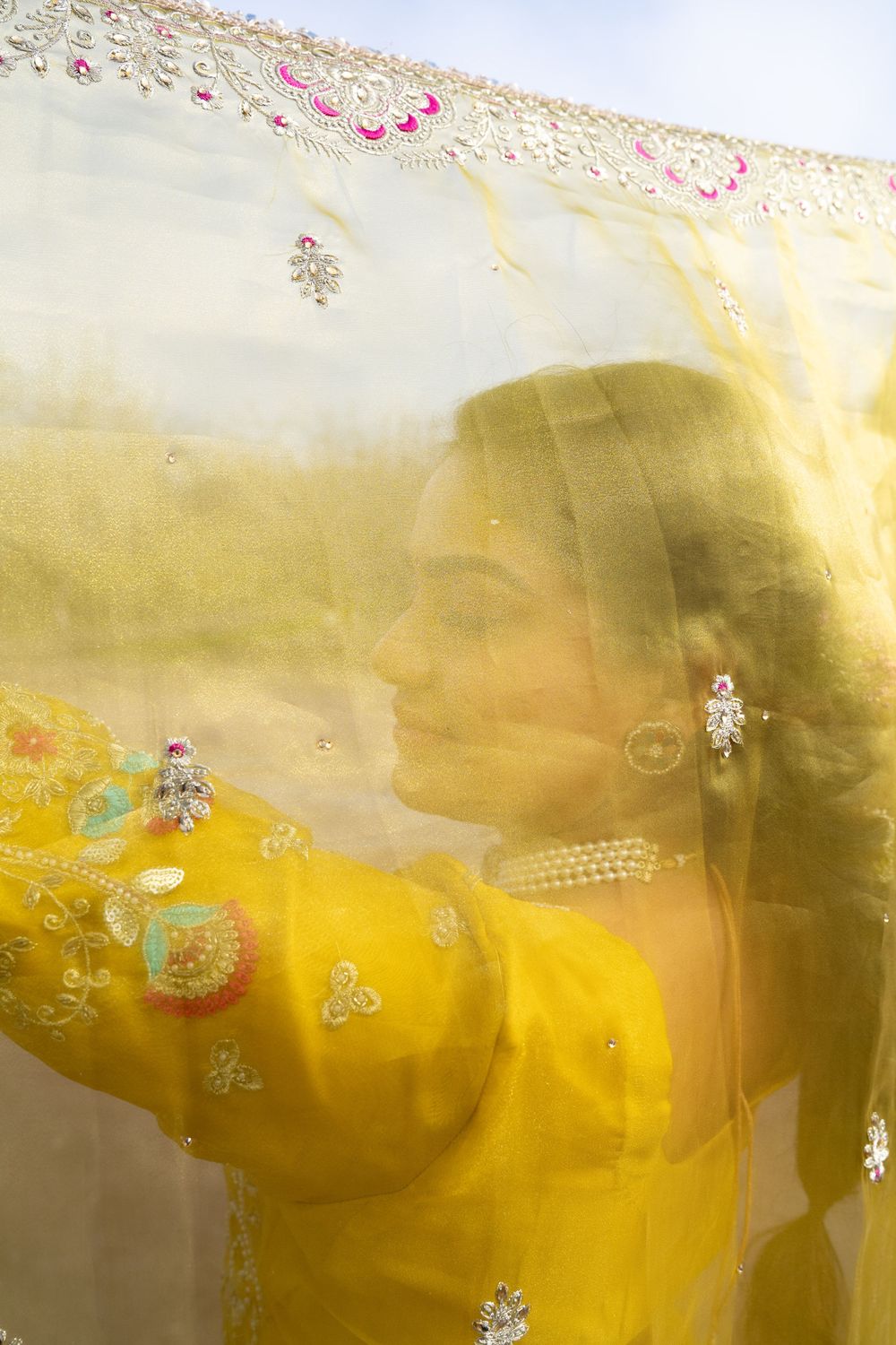 A bride in a yellow embroidered outfit poses gracefully behind a sheer golden dupatta with floral embellishments.