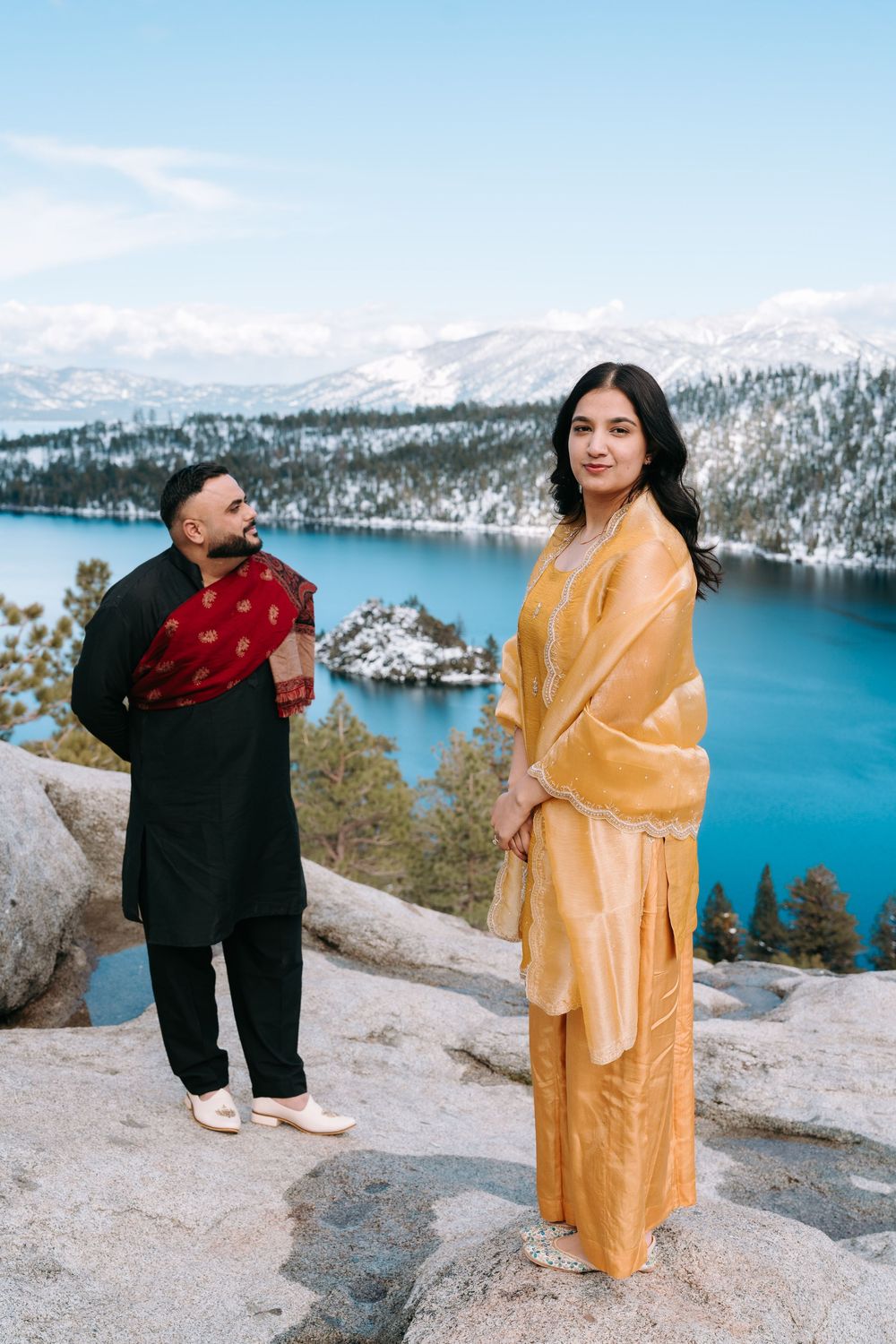 Couple in traditional Indian attire posing at Emerald Bay, Lake Tahoe, with snowy mountains and blue water.