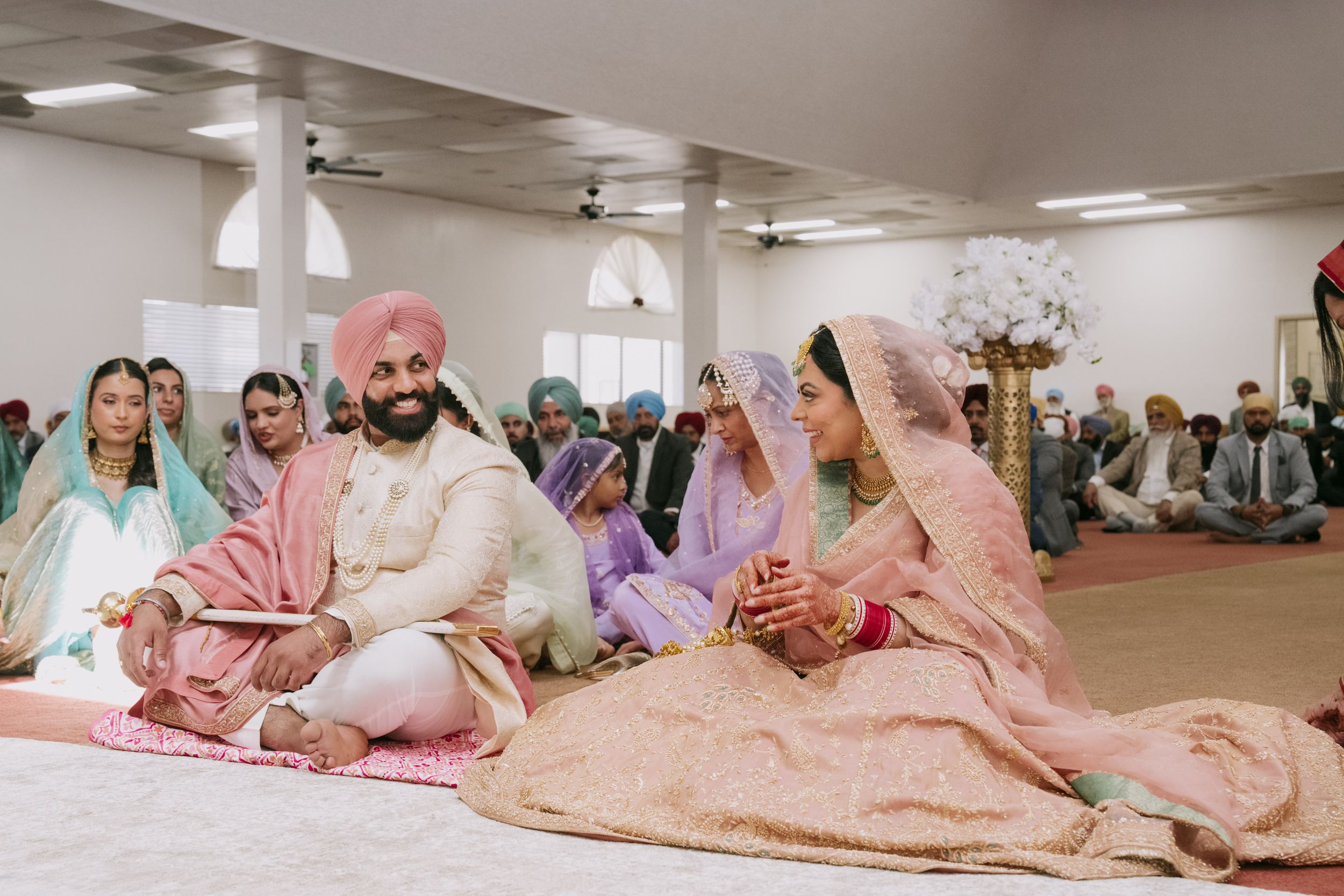 Sikh wedding ceremony with bride and groom in traditional pink attire seated on floor in a gurdwara surrounded by guests.