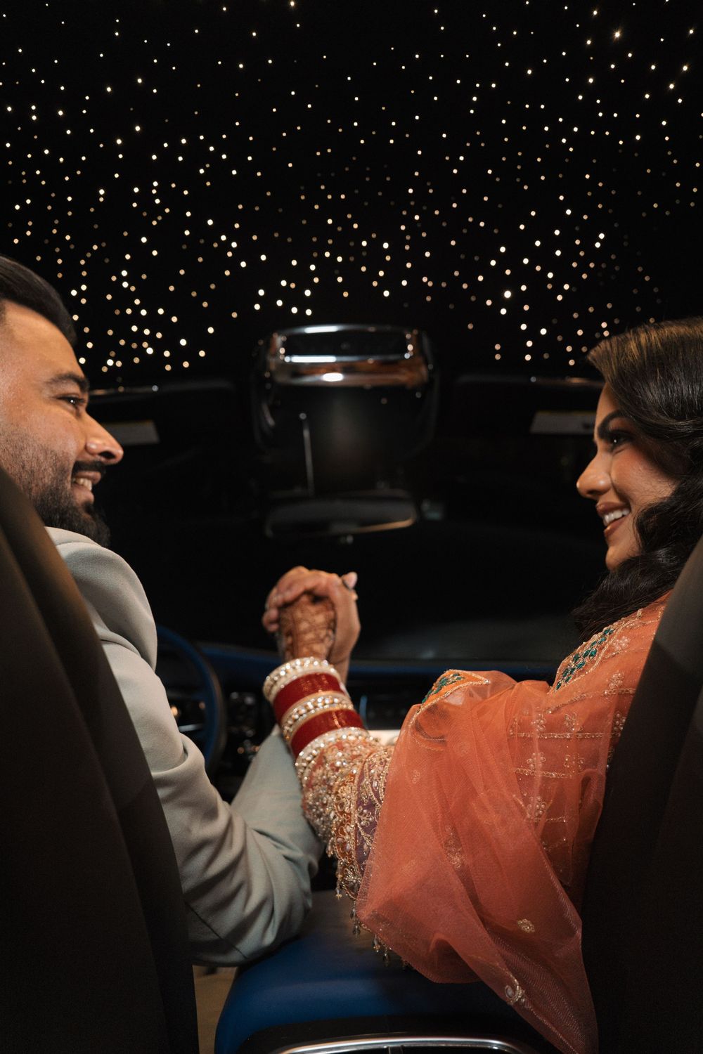 Indian couple in traditional wedding attire holding hands inside a luxury car with starlit ceiling.