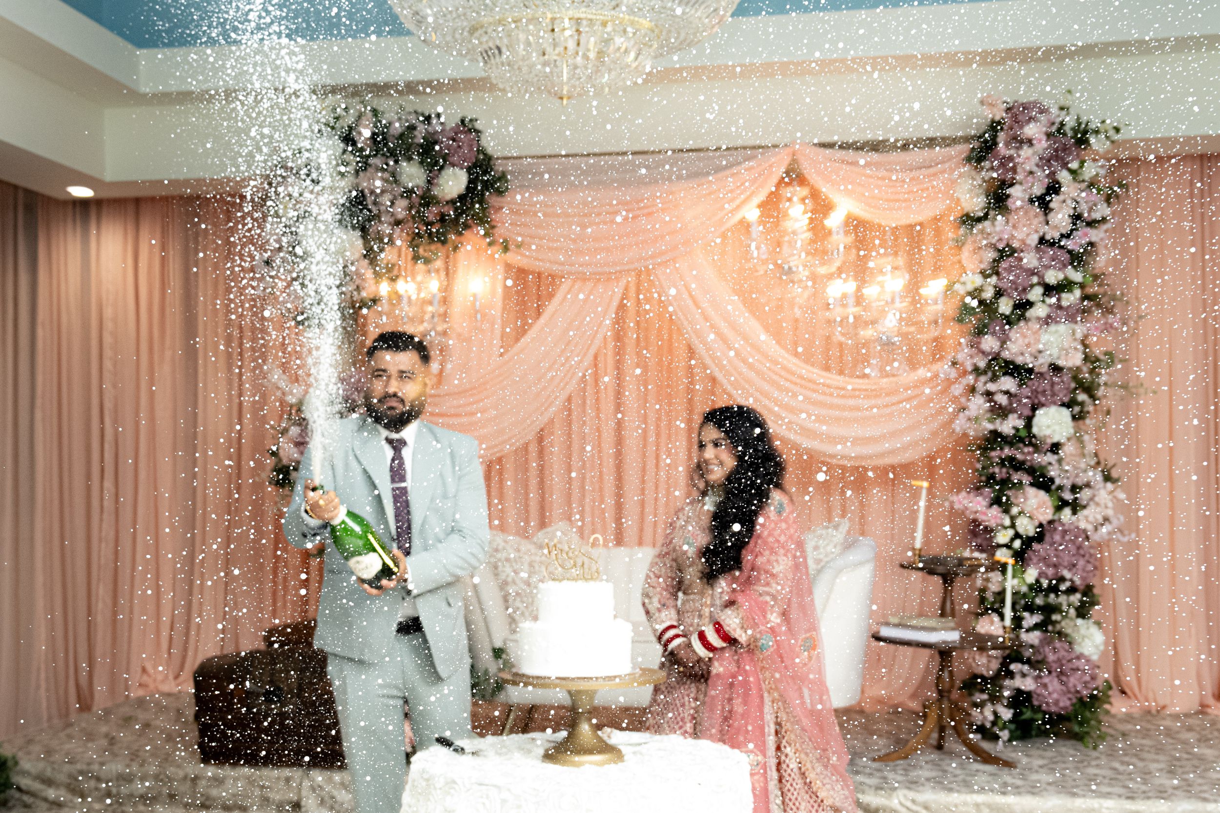 Couple celebrating at wedding reception with champagne spray near white cake, pink floral backdrop, and elegant chandelier decor.