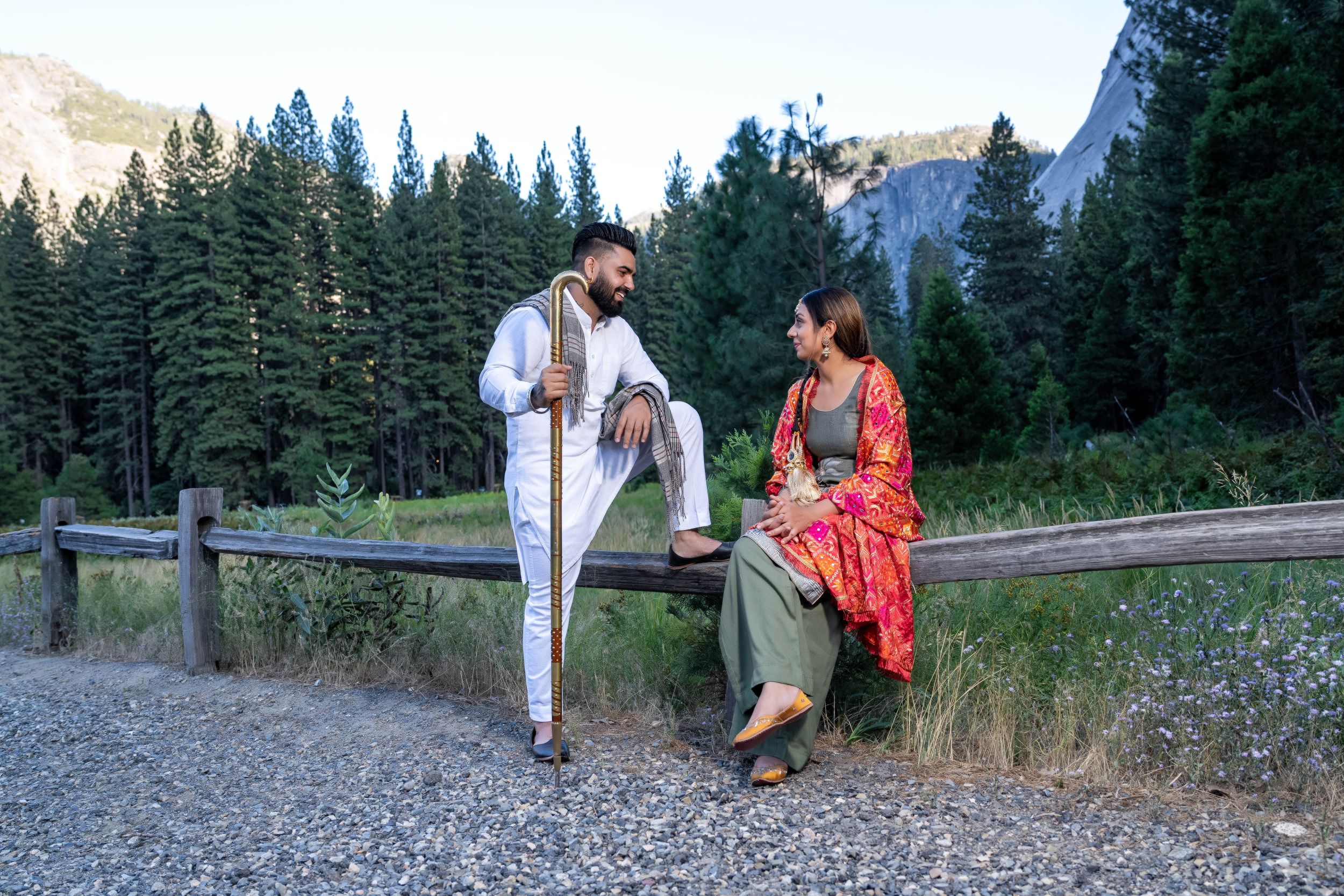 Two people in casual clothing relax on a guardrail along a mountain path surrounded by pine trees and scenic views.