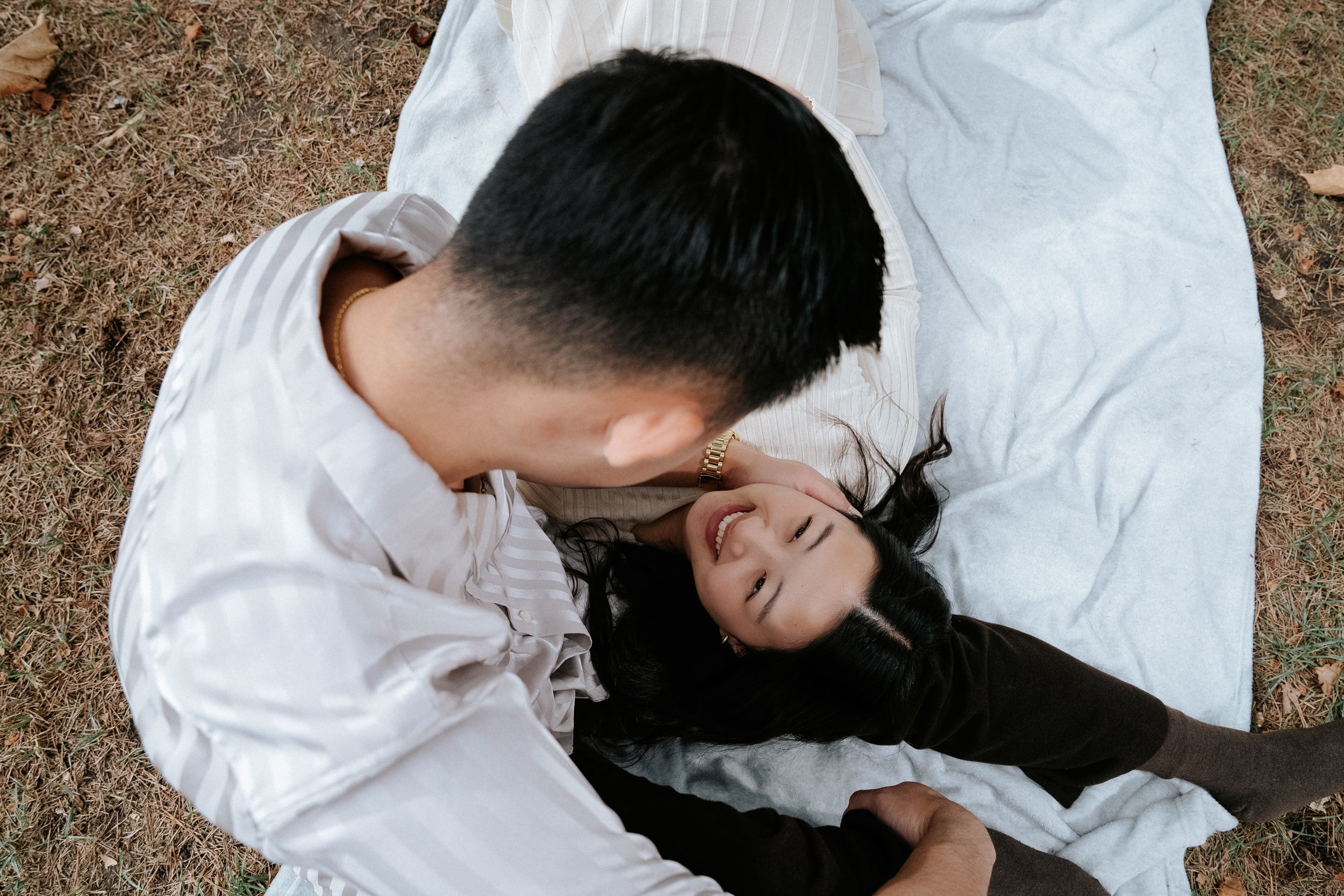 A couple in white clothing lay together on a blanket outdoors, sharing an intimate moment.
