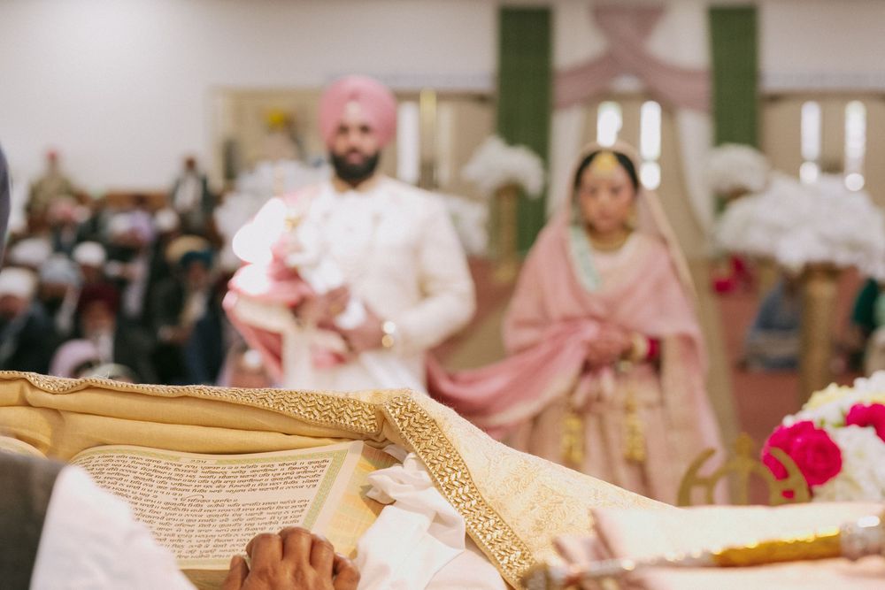 Sikh wedding ceremony with groom in pink turban and bride in blush pink dupatta standing before the Guru Granth Sahib.