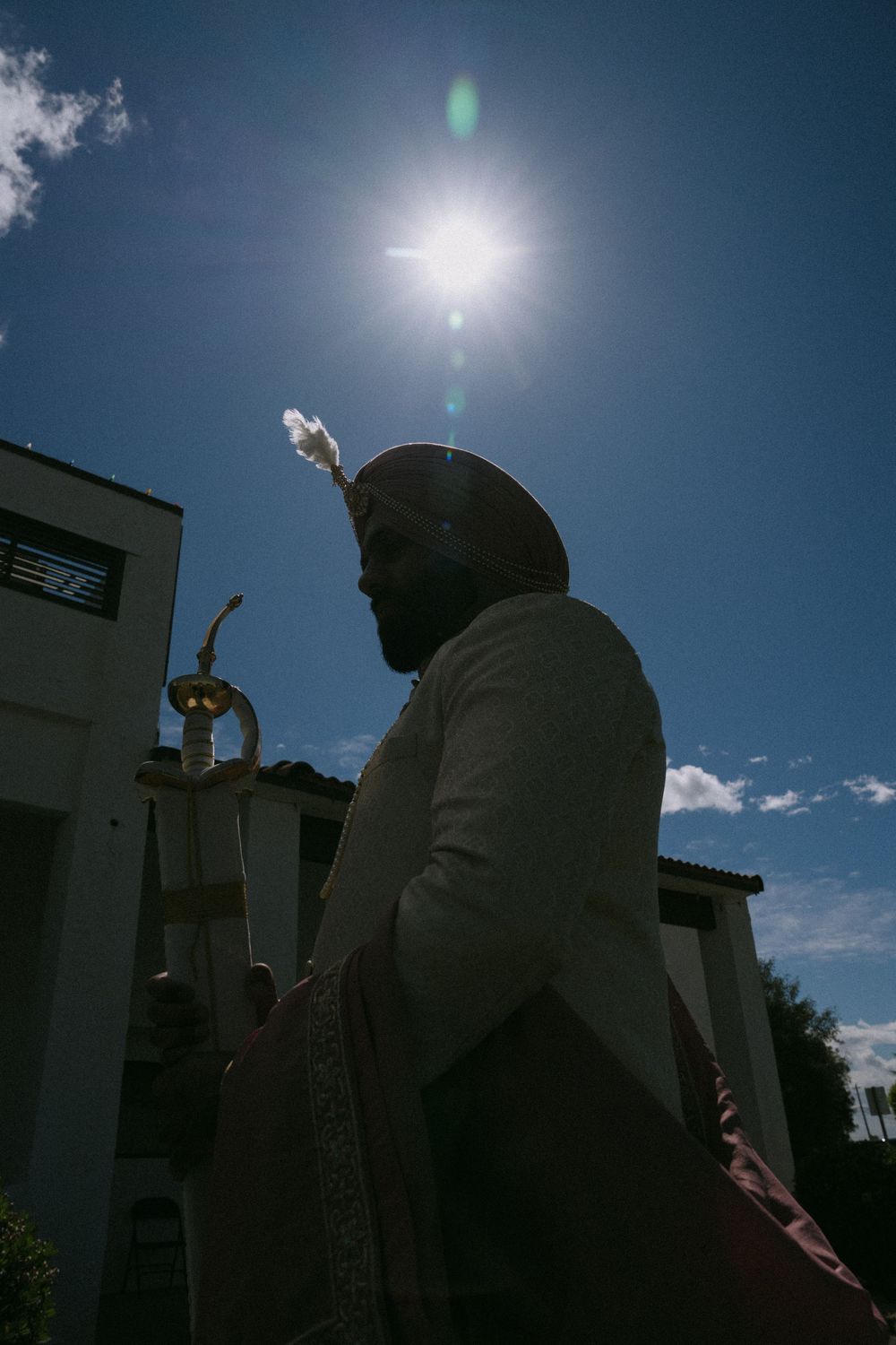 Sikh man in turban holding a ceremonial sword outdoors, backlit by bright sun against blue sky.