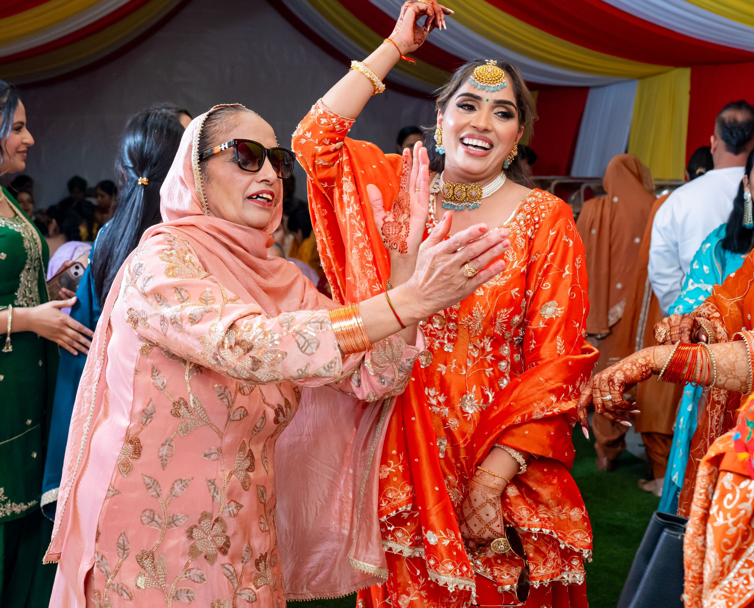 Two celebrants dance joyfully together in bright orange and pink traditional outfits during a cultural celebration.