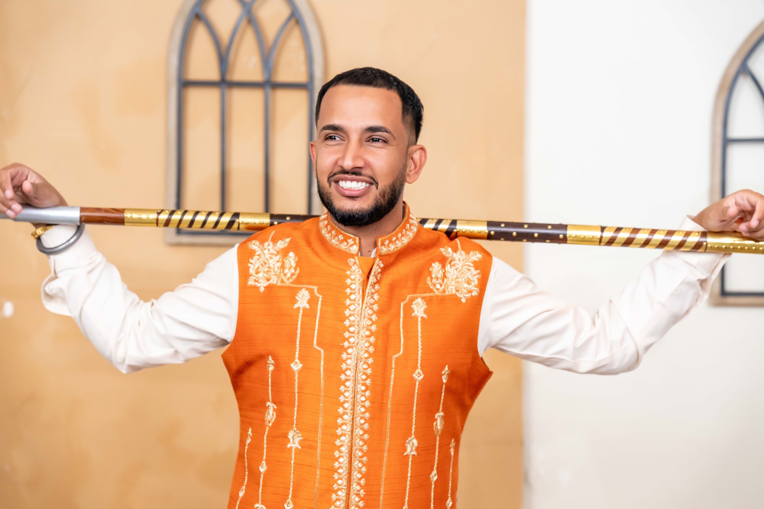 Person wearing orange embroidered vest and white shirt holds decorated stick across shoulders while smiling indoors.